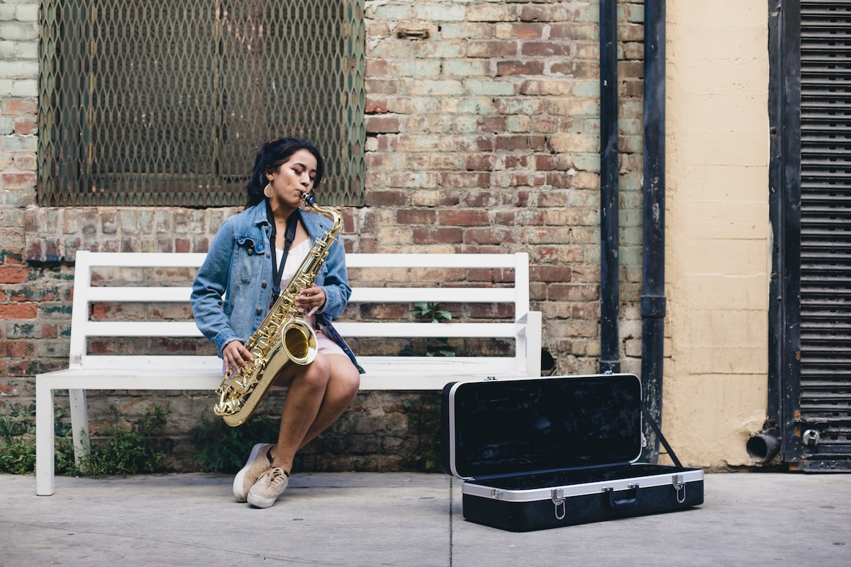 Student at the SAE tuition and audition free middle and high school playing saxophone while seated on a white bench in front of a brick wall